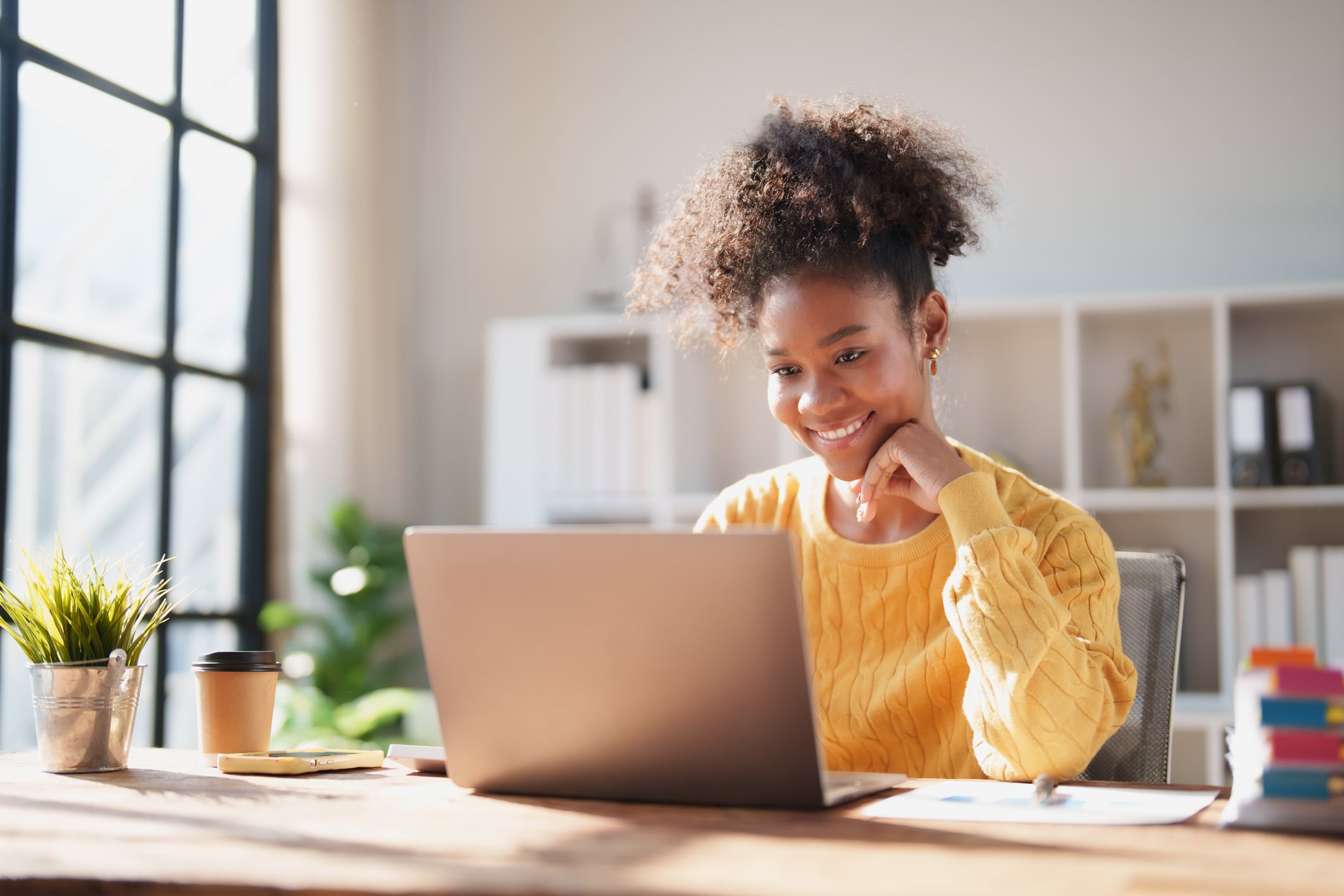 A person with curly hair in a bun and wearing a yellow sweater smiling at their laptop